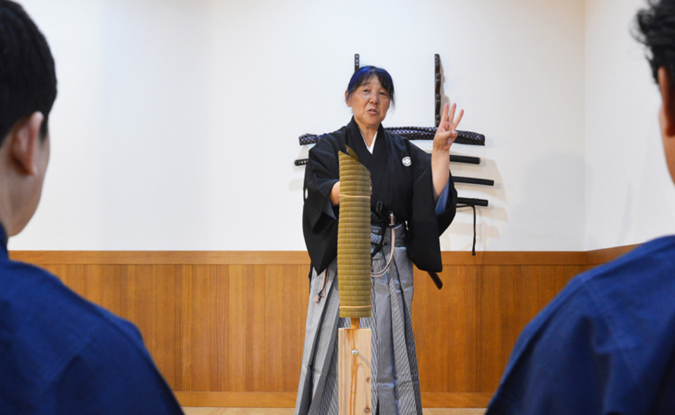 刀掛けの前で受講者に日本刀や居合について説明する講師